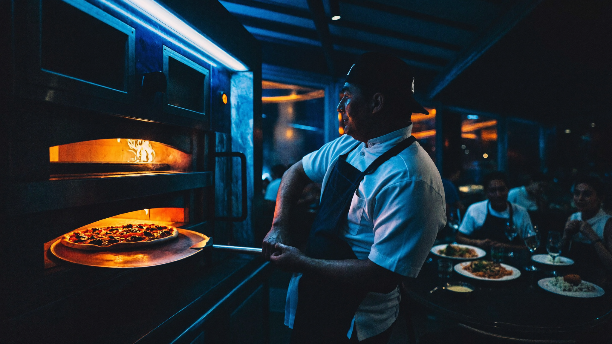 Pizzaiolo inforna una pizza nel forno a legna in una pizzeria piena di clienti